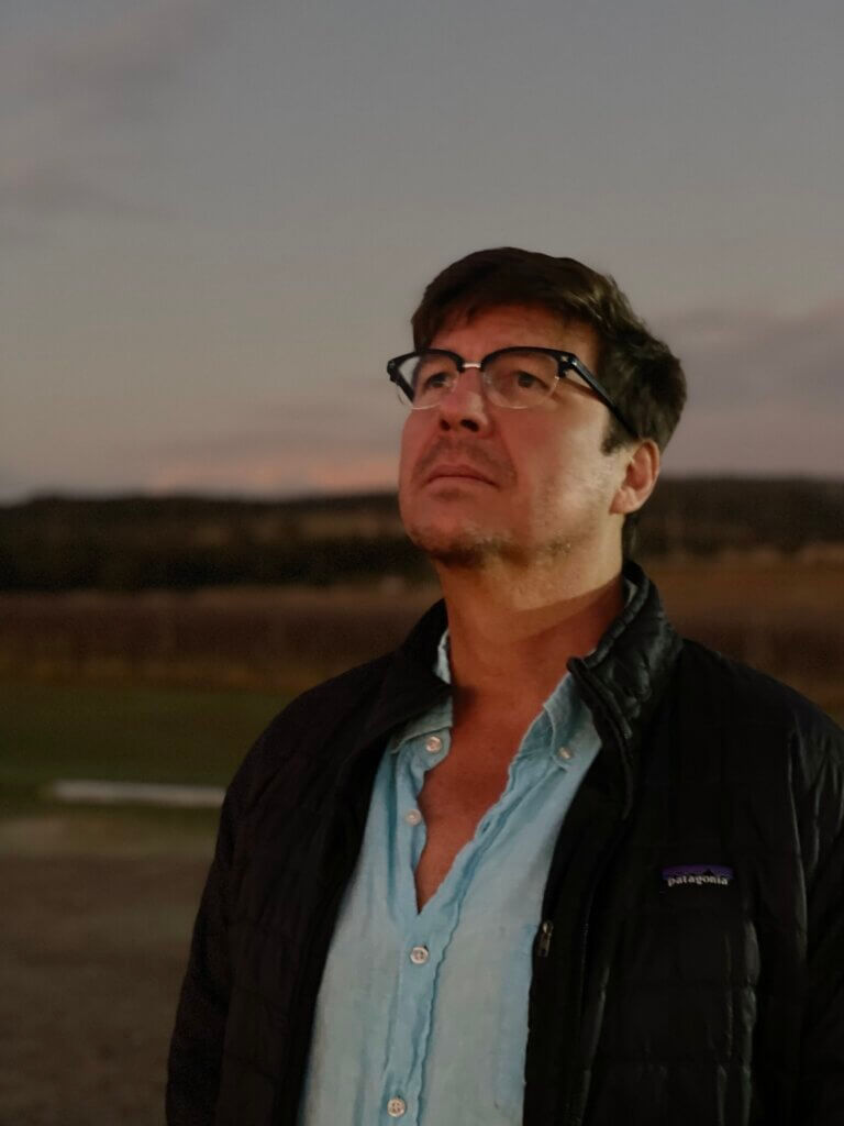 J.C. is a white male with glasses. He is wearing a light blue shirt and green jacket looking at the sky.