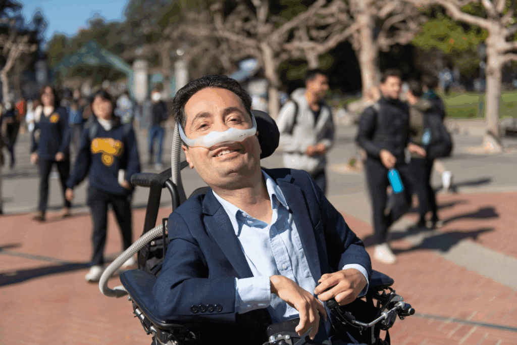 Dr. Victor Pineda smiles while seated in a power wheelchair. He is outdoors on a sunny day, with people walking in the background. He is wearing a navy blue blazer over a light blue shirt and uses a nasal mask connected to a ventilator. 