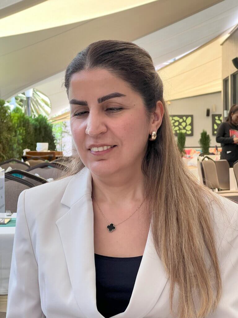 Picture of Elham Youssefian, a Middle-Eastern woman with light brown hair. She is wearing a white jacket and a black shirt, gently smiling at the camera.