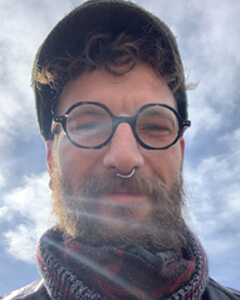 A White male in his late 30’s with a beard and hat smiling on a cloudless sunny day.