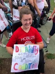 A NCIL member smiles and holds a sign that says "Disability is a natural part of the human condition"