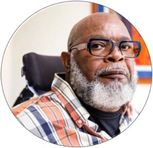 Theo Braddy Headshot: Theo is a Black man with a bald head and full beard. He is wearing blue glasses and a plaid shirt. The back of his wheelchair is visible behind him.