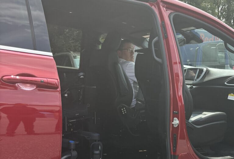 A person sitting in the driver’s seat of a red mini van. A wheelchair is visible inside the van.