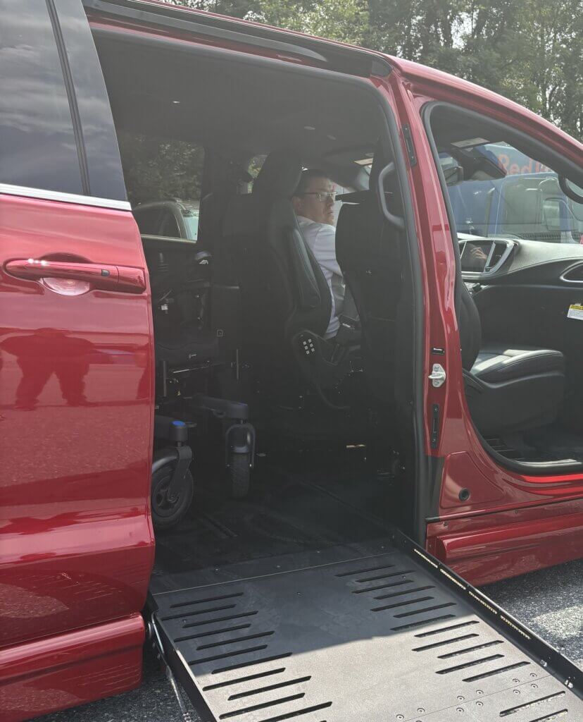 A person sitting in the driver’s seat of a red mini van with an extended ramp. A wheelchair is visible inside the van.