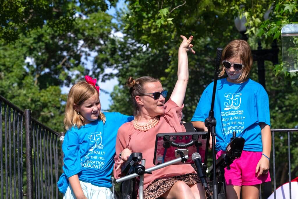 Kim Brock delivers a speech, flanked by her two daughters while on stage at the NCIL 2025 Rally at the U.S. Capitol.