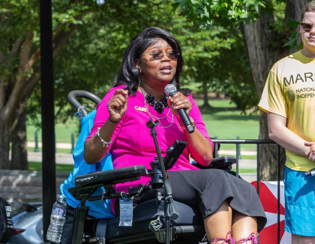 Yvette Pegues speaks into a microphone while seated in a power wheelchair during NCIL's 2025 Rally at the U.S. Capitol.