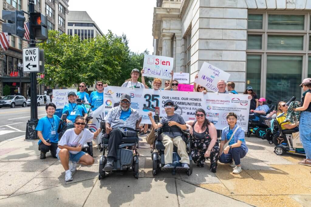 Group of 16 gathers on a sidewalk during NCIL's 2025 March to U.S. Capitol, including Theo Braddy and Victor Pineda. Participants are standing in front of a banner that says ADA35 with March and Rally sponsor logos. They are holding signs reading “Listen to Disabled Voices,” “Disability Rights are Human Rights,” and “Speak Up, Stand Out, Fight Back.”