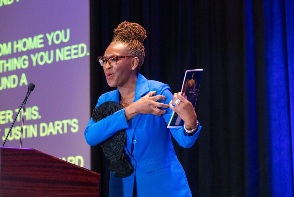 Claudia Gordon accepting the Max Starkloff Lifetime Achievement Award at #NCIL2024. Claudia is standing at a podium on stage signing “power” in American Sign Language.