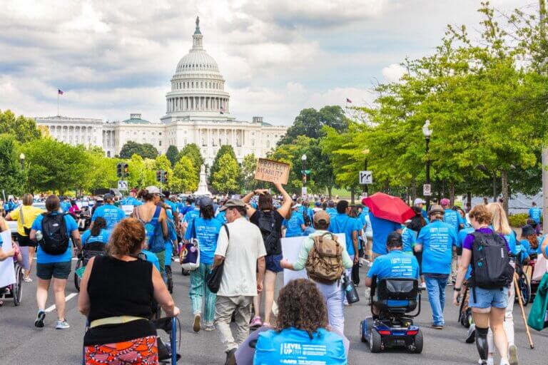 A large group of marchers, many wearing blue shirts, are seen approaching the U.S. Capitol building during NCIL's 2025 Annual Conference on Independent Living. Some participants are using wheelchairs or mobility scooters. One person is holding a sign that reads "PROTECT DISABILITY RIGHTS." The scene is vibrant with greenery and the iconic Capitol dome prominently in the background.