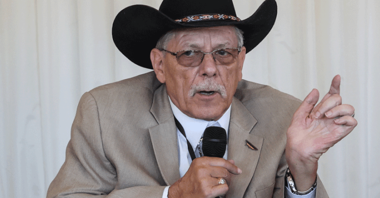 Kelly Buckland wears a cowboy hat and glasses while speaking into a microphone.