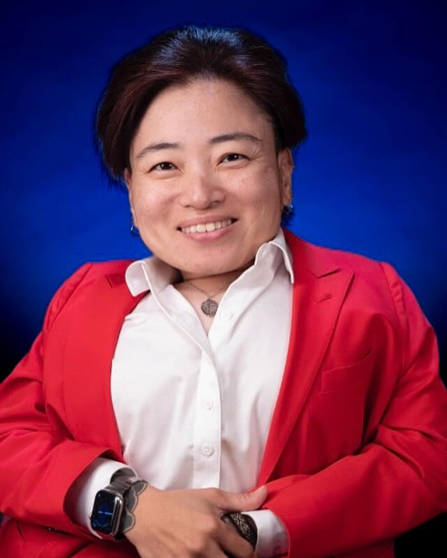 Mia Ives-Rublee wears a red blazer for a headshot against a blue studio background.