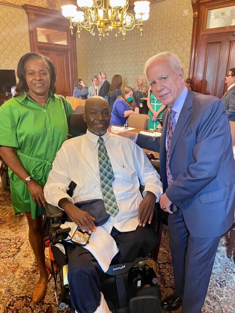 Kent Crenshaw, his wife Carmen, and Tony Coelho pose for a group photo in a formal, warmly-lit room at the White House. Kent is seated in his power wheelchair at center. Everyone is smiling.