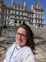 Alex, a white female in her 40's in a white ATI t-shirt in front of the NY State capital.