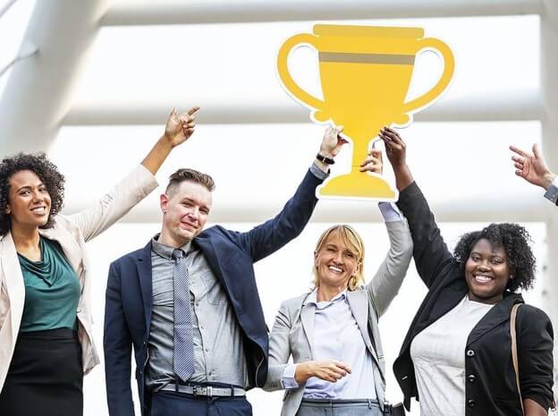 Photograph of a diverse group of four business professionals smiling and holding up a large yellow trophy cutout, symbolizing achievement or success. The group is dressed in business attire, standing under a bright, modern architectural structure, highlighting teamwork and celebration.