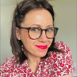 Courtney Williams, a woman with short brown hair wearing tortoiseshell glasses and bright red lipstick, smiling slightly. She is dressed in a white shirt with a red floral pattern and is positioned against a plain light-colored background. 