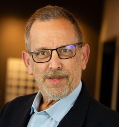 Jim House, a middle-aged man wearing glasses, a light blue shirt, and a dark blazer, posing indoors with a blurred background. The setting suggests a professional or formal environment, with warm lighting highlighting the subject's face.