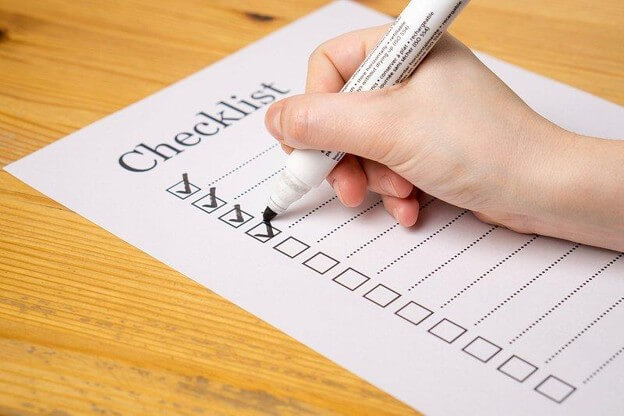 Photograph of a hand holding a white marker and marking checkboxes on a printed checklist sheet placed on a wooden surface. The checklist has multiple empty checkboxes with some already checked, indicating progress or task completion.