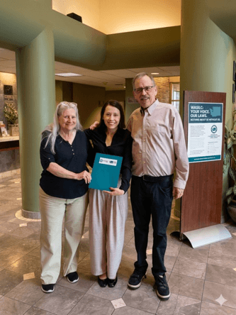 Photograph of three people standing indoors in a lobby area, smiling and posing for the camera. One person in the center holds a blue folder, while a signboard nearby displays text about "WASILC" and encourages voicing opinions on laws.